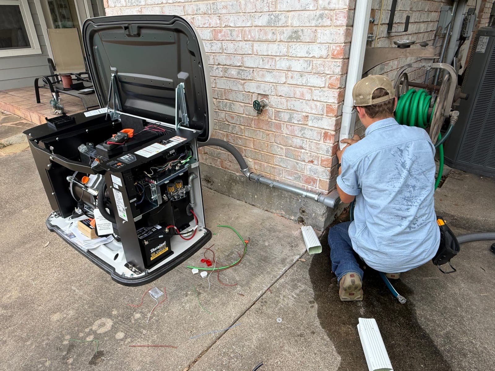 An Armored Power Systems technician kneeling at an open home standby generator, working on the connections at the base of the enclosure.